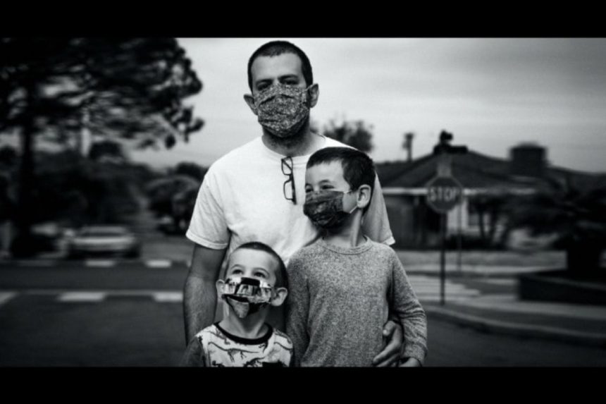Man and his sons stand together, masks on, in a somber black and white toned photo